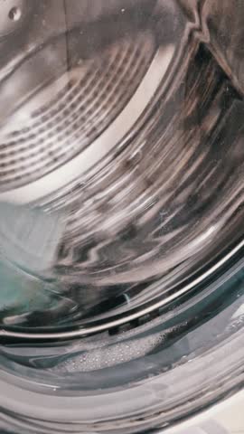 Vertical, close up shot from inside a front-load washing machine drum. Light blue, green, and red towels are tumbling in water and soap suds during a vigorous and colorful wash cycle.