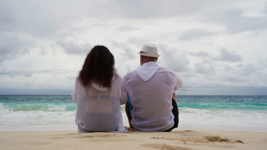 Rear view of couple in white clothing sitting side by side on beach sand, man in white hat, enjoying peaceful ocean view under dramatic cloudy sky. A woman puts on a hat