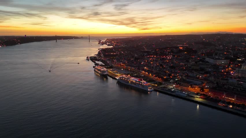 A drone view of Lisbon, Portugal, at sunset with illuminated cruise ships along the Tagus River and the 25 de Abril Bridge on the horizon
