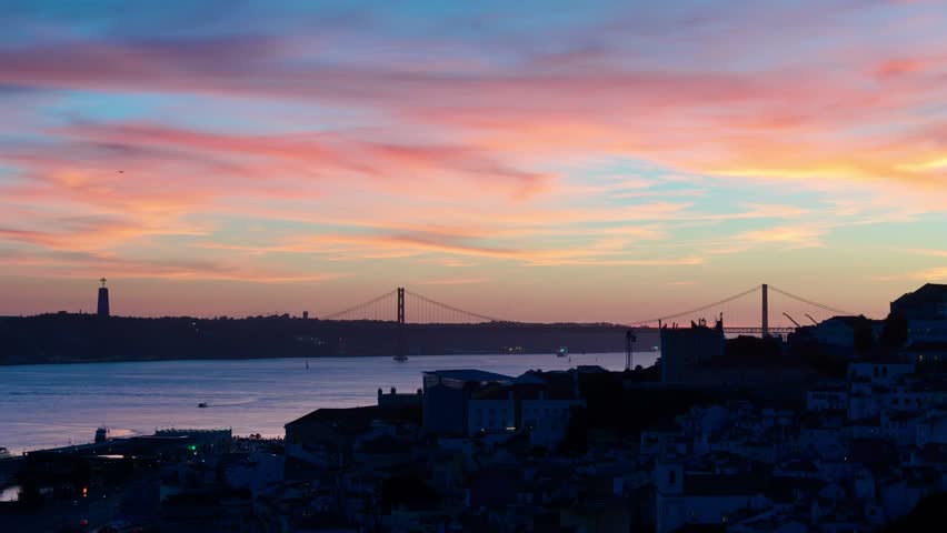 An aerial view of Lisbon, Portugal at sunset, with the 25 de Abril Bridge and Cristo Rei monument silhouetted against dusk over the Tagus River