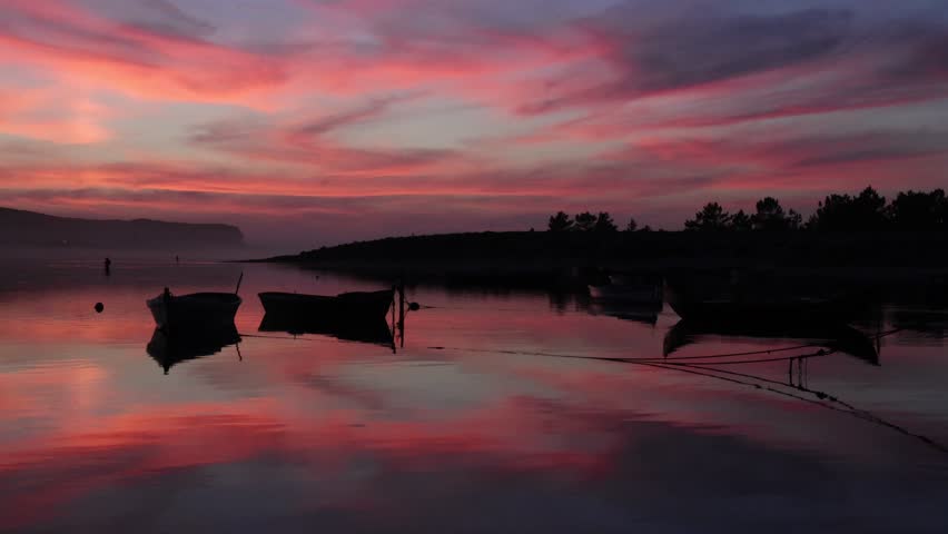 A scenic view of small fishing boats floating on still water under a vivid pink and orange sunset sky, Obidos Lagoon, Portugal, reflecting beauty