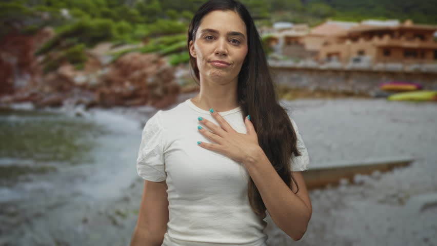 Woman with hand on chest at seaside building by the shore, smiling with turquoise nails and long hair in white shirt, standing outdoors; gratitude.