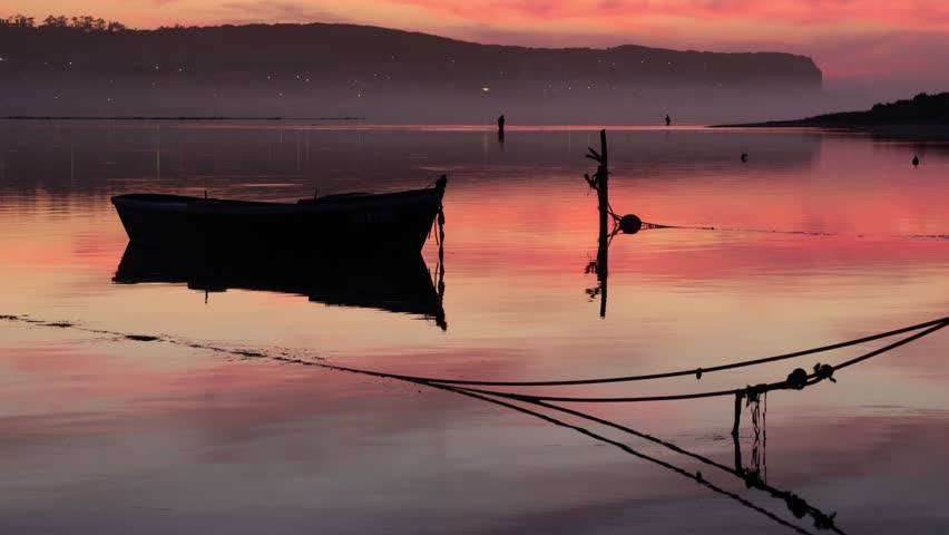 A solitary boat anchored on Obidos Lagoon, Portugal, under a glowing red and purple dusk sky with reflections shimmering on the calm water surface