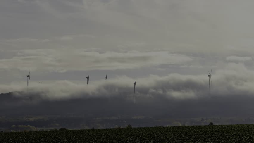 A timelapse of landscape with wind turbines emerging through a blanket of clouds over lush green farmland
