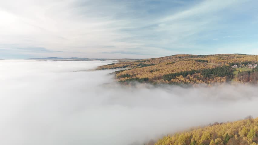 A drone aerial view of a hilly autumn forest landscape shrouded in fog and low clouds under blue, white sky in the Czech Republic