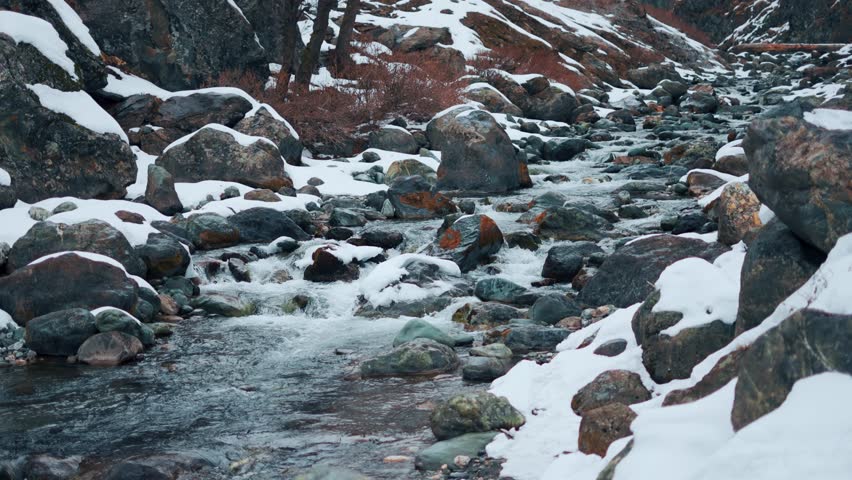 4K shot of slow flowing river and rocks with snow on sides at Manali, Himachal Pradesh, India. Natural winter landscape. Serene river stream in snowy Himalaya mountains. 