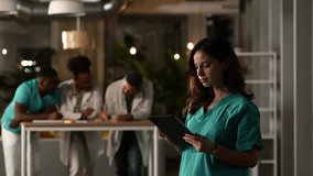 Young female medical professional in scrubs using a tablet computer. She smiles at the camera while her diverse team works in the background - Powered by Shutterstock - Get 15% off with code: PIKWIZARD15