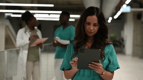 Young smiling nurse in scrubs using a digital tablet in a hospital hallway. Diverse medical team in the background discussing patient care - Powered by Shutterstock - Get 15% off with code: PIKWIZARD15