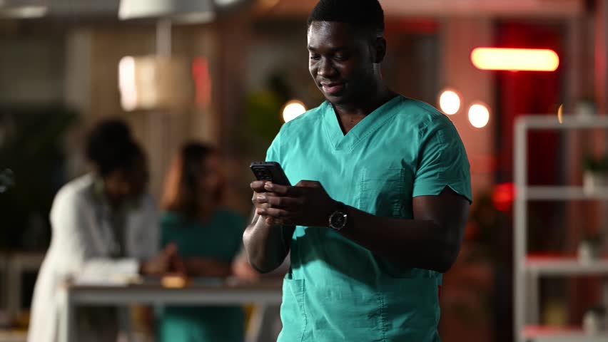 Anxious male nurse in scrubs looking at smartphone and answering a stressful phone call. Agitated medical assistant working late at night