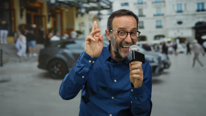 Man speaking into microphone outdoors in urban setting with blurred background of buildings and people in casual attire.