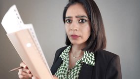 A concerned South Asian woman in a blazer holds a file and pen, looking at the camera with astonished amazed face expression against gray wall - Powered by Shutterstock - Get 15% off with code: PIKWIZARD15