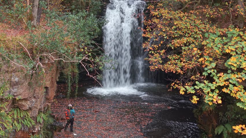 Aerial view of a lone hiker admiring a cascading waterfall in autumn, Wales.