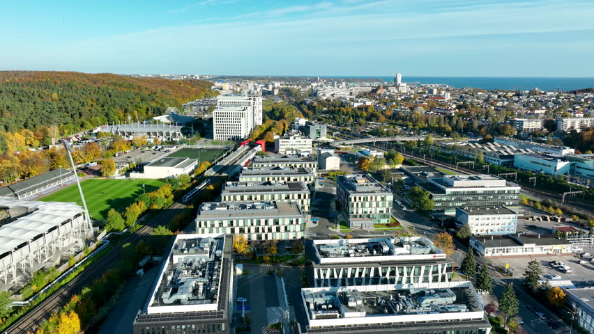 Aerial panorama of Gdynia with modern offices, stadium, and sea horizon visible in the distance on a sunny day.
