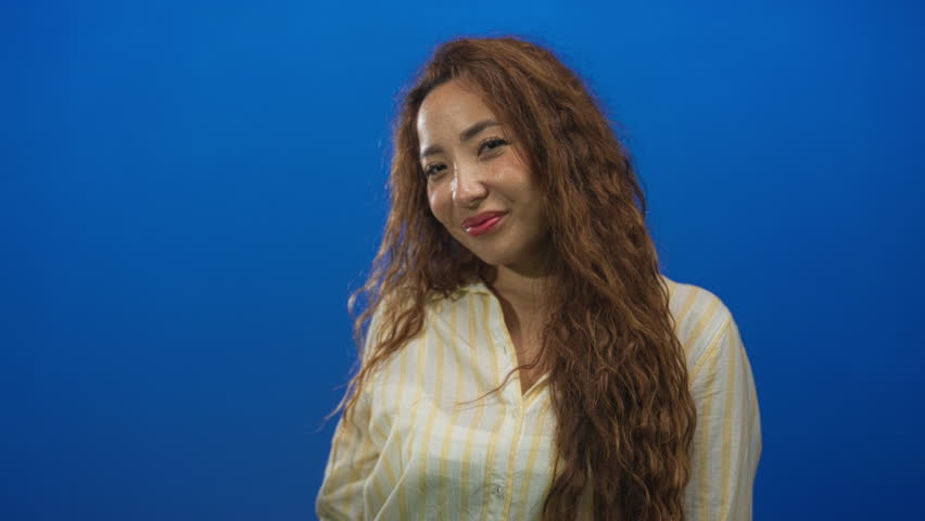Woman pointing and saluting with her hand, smiling with long curly hair and striped shirt in blue studio; confidence.