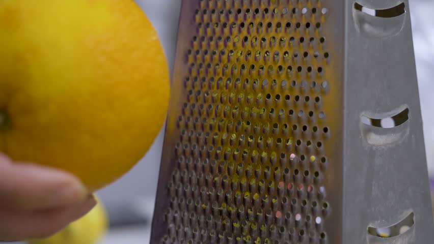Female chef hand rubs orange against metal grater into fine pieces. Zest gathers for cookie mixture. Effort, timing, and recipe accuracy at kitchen. Shredder utensil used to grate or shred foods