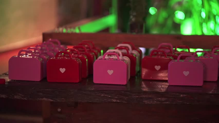 A close-up of cute mini suitcase wedding favors in pink and red with a heart detail, displayed on a rustic wooden table at a reception.