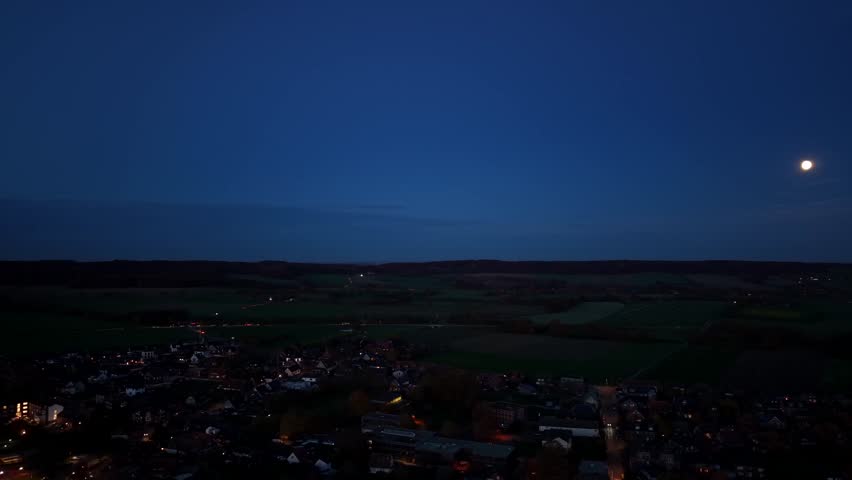 Full moon at blue dusk sky in american town. Lighting houses and streets in USA. Aerial lateral wide shot. Rural landscape with hill farm fields at night. Panorama view.