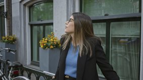 Woman standing on european city street with glasses and brunette hair, wearing blue sweater and black coat, smiling near bike rack with flowers by window. - Powered by Shutterstock - Get 15% off with code: PIKWIZARD15
