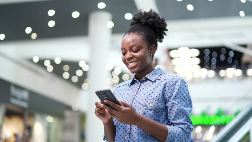 Young smiling african american woman shopper using mobile phone standing in shopping center mall. Happy joyful black woman customer browsing web, chatting online, typing using smartphone in store