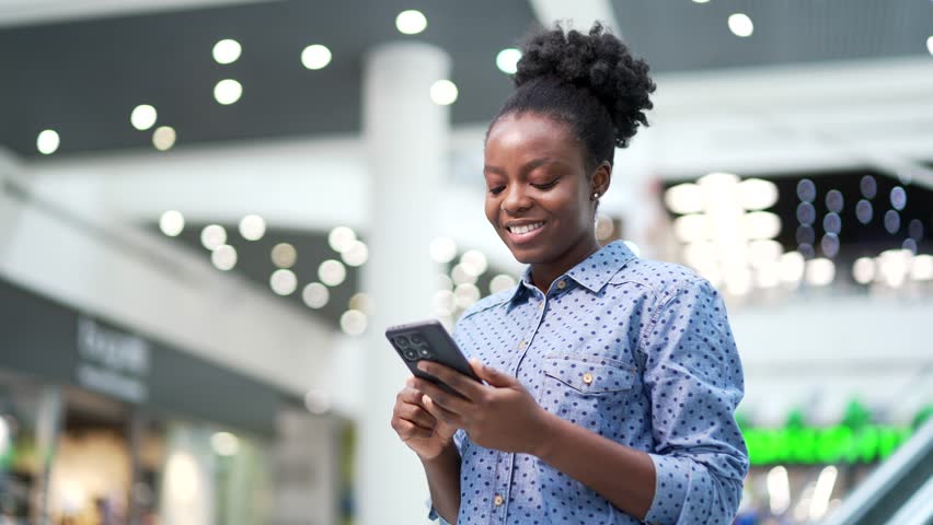 Young smiling african american woman shopper using mobile phone standing in shopping center mall. Happy joyful black woman customer browsing web, chatting online, typing using smartphone in store