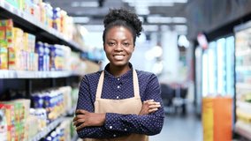 Portrait of a smiling African American female supermarket worker standing with crossed arms between rows of shelves in market grocery store. Happy sales manager or owner in apron looking at camera - Powered by Shutterstock - Get 15% off with code: PIKWIZARD15