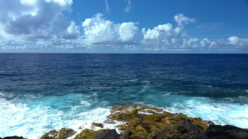 The Hawaiian coastline unfolds in vivid color as dense green rainforest stretches toward the deep blue Pacific, where waves carve ancient volcanic cliffs under dramatic island skies.
