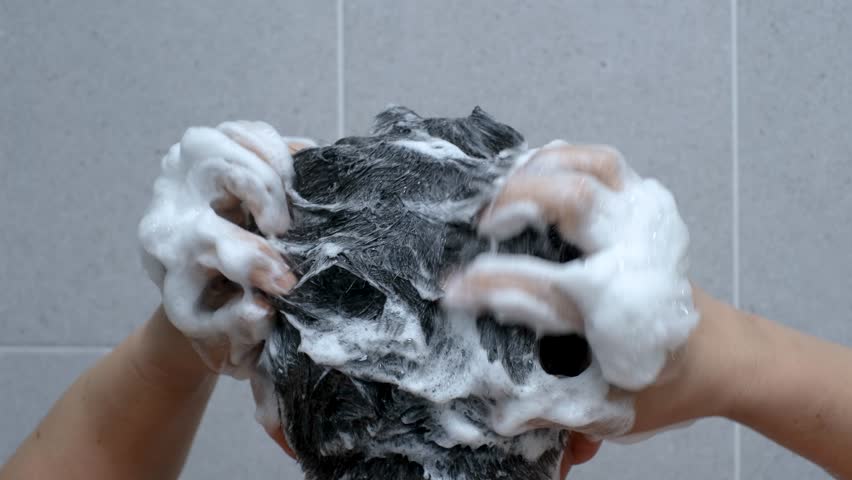 Male hands wash their hair with shampoo and foam on gray tile background close-up.