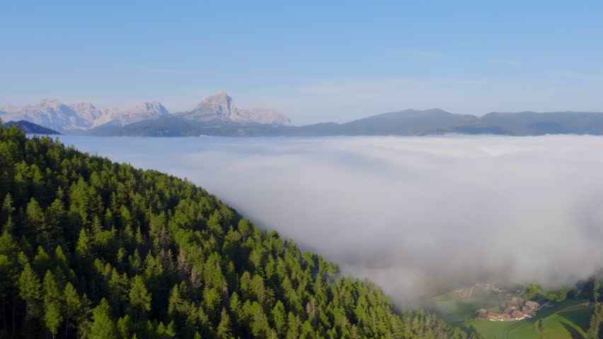 Drone flight over the Dolomites in morning fog. The camera moves gently downward to the right, revealing forests, mist, and sunlit mountain peaks in a vast landscape.