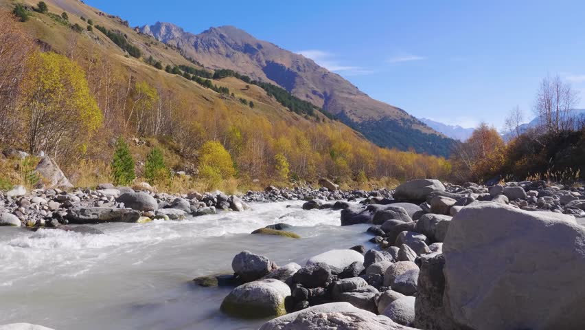 A scenic view of a mountain river, possibly the Baksan River, flowing through a rocky gorge in the North Caucasus region of Russia. The surrounding mountains are covered in autumn foliage, with a mix