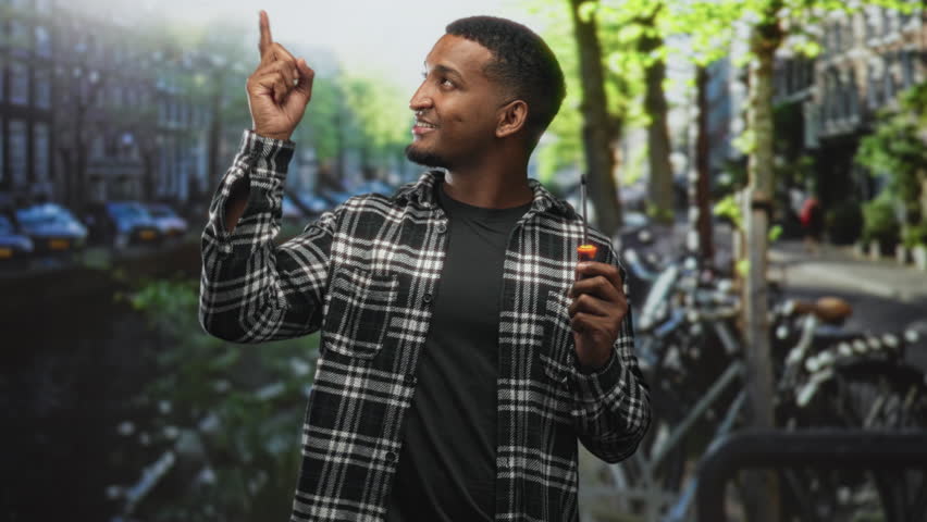 Man holding screwdriver points finger up on street beside canal and parked bicycles, smiling and making a pointing gesture; confidence resourcefulness.