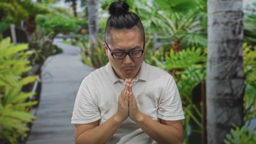 Man with palms together praying on a wooden forest walkway wearing white polo and glasses; serenity meditation.