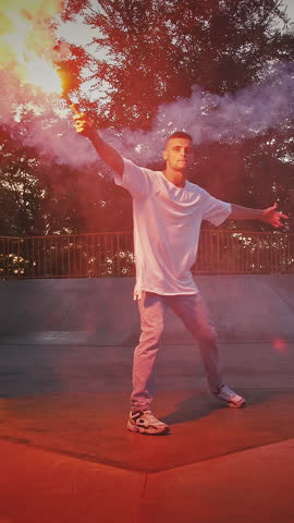 Young male in casual outfit is holding glowing red signal flare while performing street dance on pump track of rollerdrome in the evening. Hip hop and breakdance technique. Slow motion