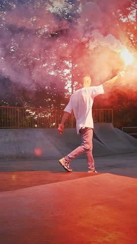Young guy in casual clothes is holding glowing red signal flare, jumping and spinning while performing street dance on pump track of skatepark. Hip hop and breakdance technique. Evening, slow motion