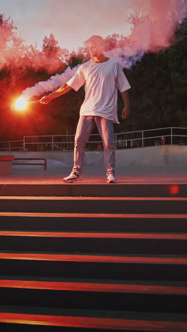 Young fellow in casual outfit is holding glowing red signal flare and going to perform street dance standing on stairs of rollerdrome in the evening. Hip hop and breakdance technique. Slow motion