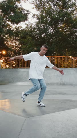 Young man in casual clothes is smiling, jumping and spinning while performing street dance on pump track of skatepark. Hip hop and breakdance technique. Early morning, sunrise