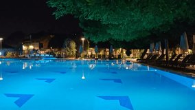 Sun loungers and umbrellas are placed among the trees next to the illuminated pool. Nearby there are flower beds, bridges, lanterns and buildings. People are walking. Warm autumn evening and twilight - Powered by Shutterstock - Get 15% off with code: PIKWIZARD15