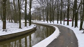 The partially ice-covered canal runs through the park among spruce and poplars trees. The banks are concreted Snow covers the ground. Nearby there is a tiled path, lampposts, parked cars and buildings - Powered by Shutterstock - Get 15% off with code: PIKWIZARD15