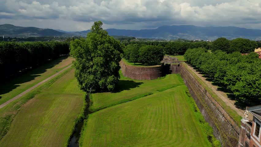 Aerial drone footage approaching the walls of Lucca in Tuscany, Italy, revealing both the fortified structure and the surrounding town with trees, rooftops, and open parkland in summer.