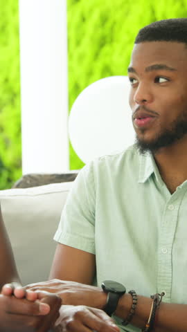 Vertical video: African American couple receiving small gift exchanging larger box on sofa at patio. Romance, celebration, joy, intimacy, decor, happiness, vertical video