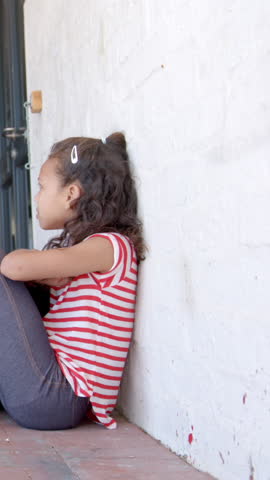 Female child curling knees on brick walkway by locked padlock, hair clip, striped tee, copy space. Vulnerability, resilience, childhood, confinement, outdoor, minimalism, innocence