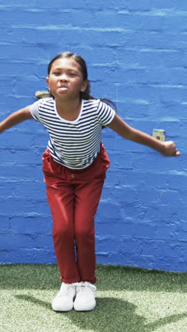 Vertical video: Girl jumping after bending knees and swinging arms playfully by blue wall on turf. Children, play, outdoor, activity, lifestyle, cheerful, vertical video