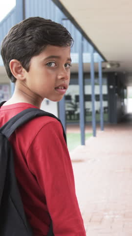 Vertical video: Asian preteen boy walking to class with backpack, glancing and smiling, copy space. Children, school corridor, urban environment, candid moment, youthful, education, vertical video