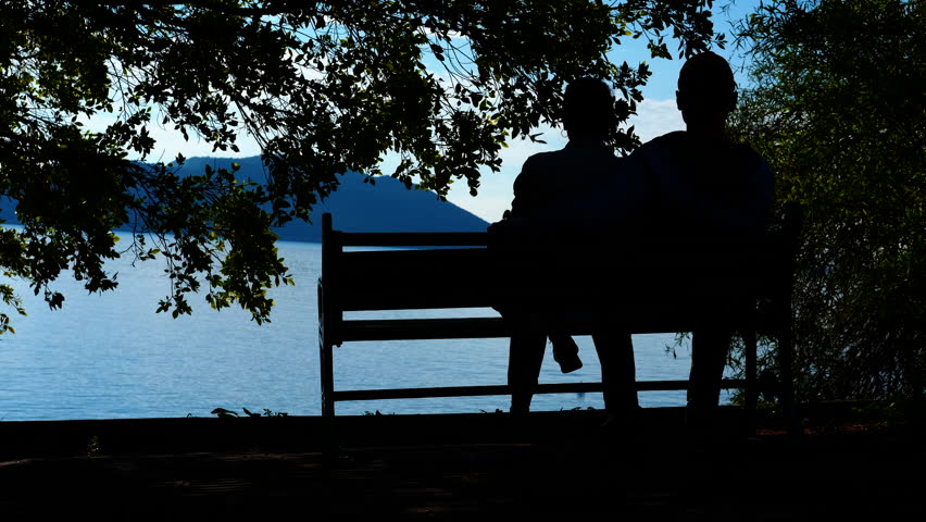 Couple silhouette relaxing on bench by lake. Silhouetted couple embracing closely on wooden bench, sharing intimate moment with tranquil lake landscape during golden evening light