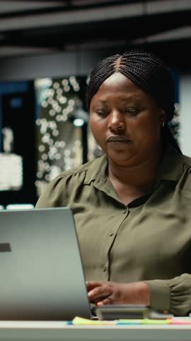 Vertical Video African american woman closing her laptop and office lamp, preparing to leave the building, going home at midnight. Employee working hard after hours, alone in dark space. Camera B.