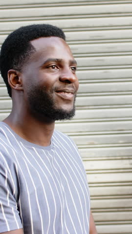 Vertical video: African American man turning to camera, crossing arms and laughing against shutter. Casual, urban, candid, expressive, upbeat, modern, vertical video