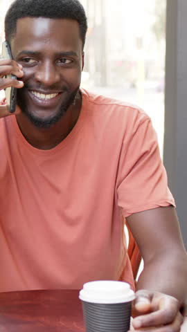 Vertical video: African American man at cafe answering smartphone call, pointing outside for view. Casual, professional, lifestyle, warm, candid, natural, vertical video