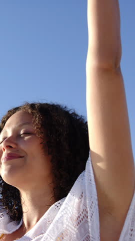 Vertical video: Woman wearing lace coverup raising arms tilting head under blue sky, copy space. Freedom, serenity, relaxation, wellness, mindfulness, openness, vertical video