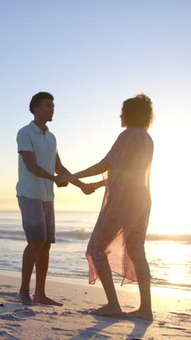 Vertical video: Sunset inspiring couple dancing on shore then man dancing solo and woman running. Romance, serenity, vitality, leisure, nature, harmony, vertical video