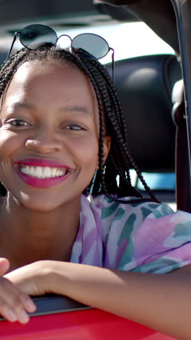 Vertical video: African American woman in red car sliding sunglasses down to block sun, copy space. Lifestyle, leisure, fashion, travel, outdoor, vibrant, vertical video