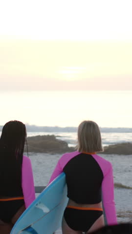 Walking diverse female surfers carrying light blue surfboard along sandy beach at sunset. Beach, surfing, adventure, outdoor, vibrant, friendship, leisure
